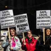 Three women at a prochoice rally holding signs that say "Safe, legal abortion on demand. No apologies!" and "Abortion is a right. No Going Back."