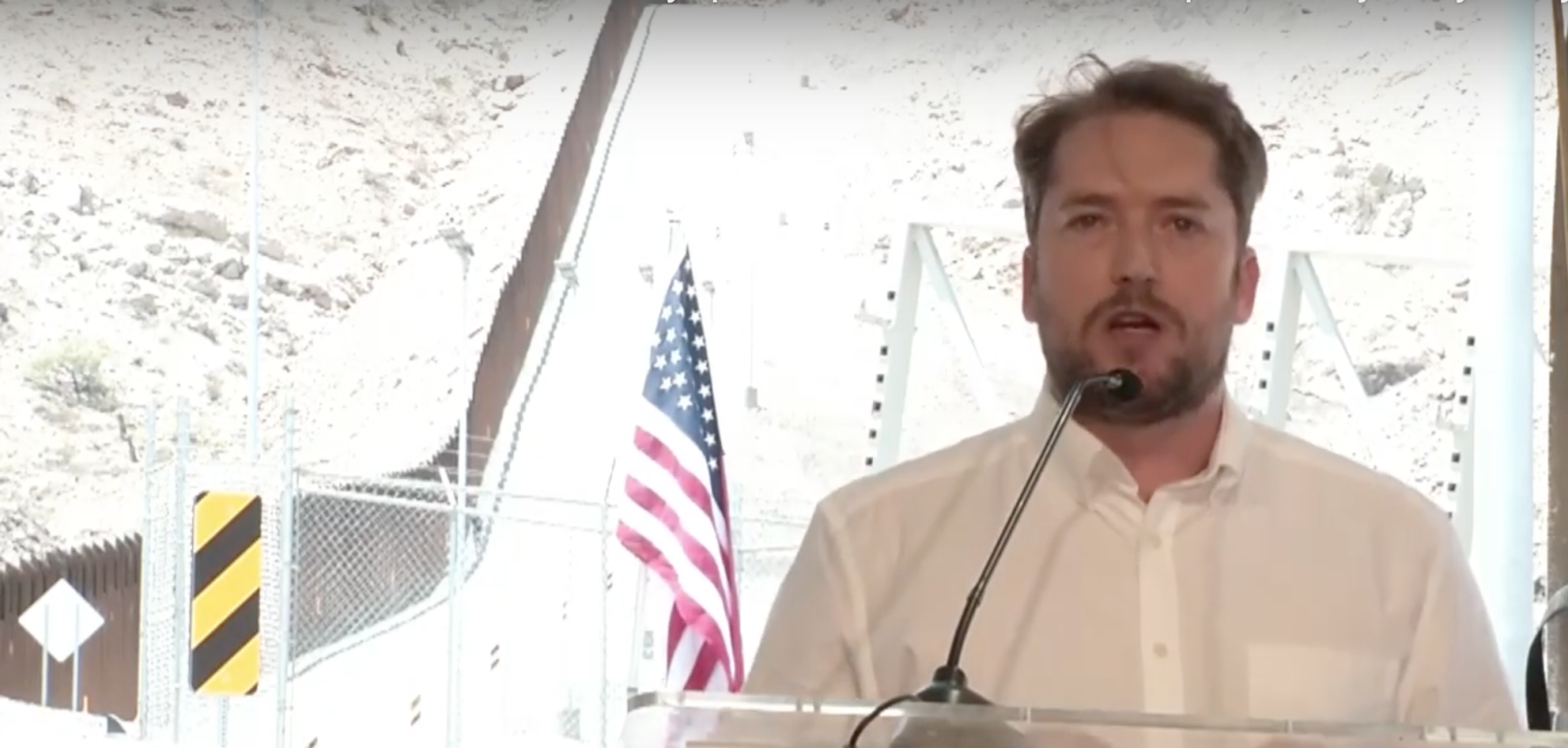 Darren Beattie delivers speech in NM with U.S.-Mexico border wall construction behind him. 