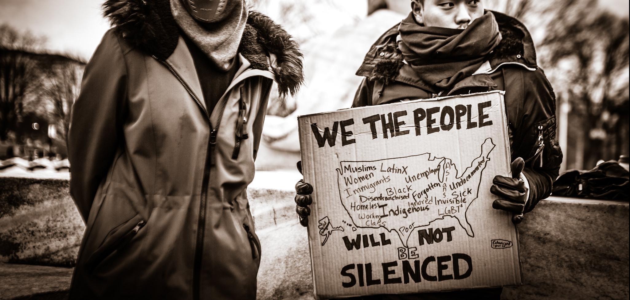 Two men in winter clothes, one holding a sign that reads "We the people will not be silenced"