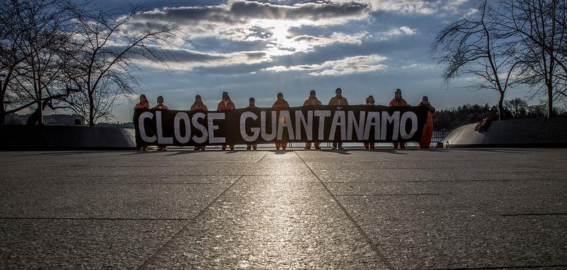 A row of people in orange jumpsuits holding a sign that says "close Guantanamo"
