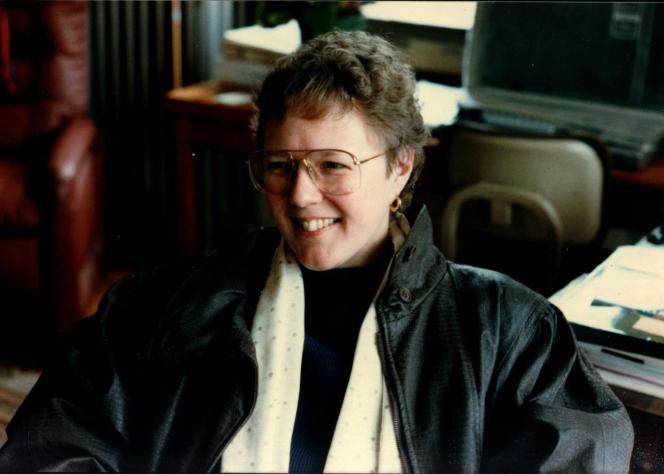 A white woman with big glasses and short hair, wearing a black jacket and a white scarf. She is sitting in a chair in an office.