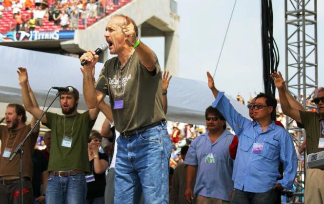 A tall white man in a green t-shirt and jeans screaming into a mike with people standing behind him.