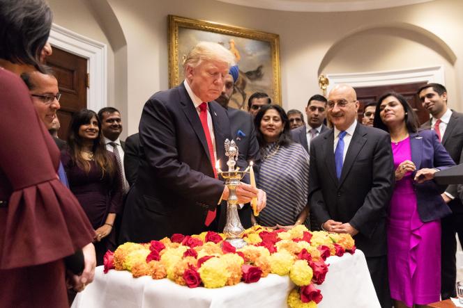 Trump using a candle to light a lamp that's set on a table of flowers with people surrounding him.