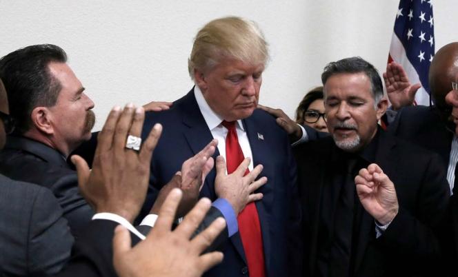 President Trump with men standing around him, all of their hands are on him and their eyes are closed in prayer
