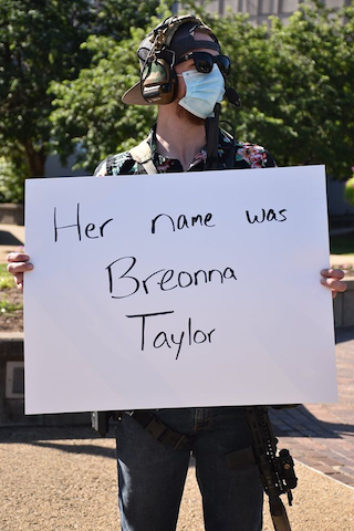 White man wearing Hawaiian shirt, firearm and tactical gear holding "Her name is Breonna Taylor"