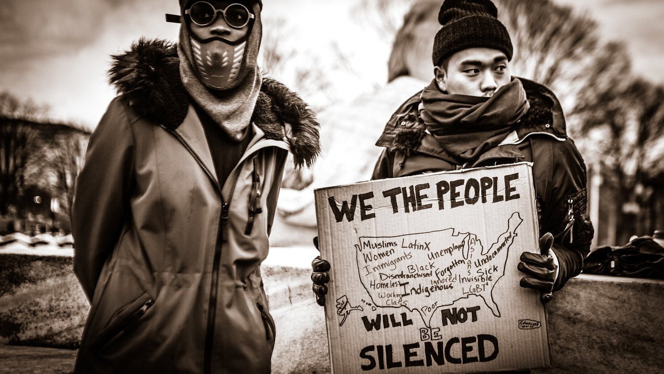 Two men in winter clothes, one holding a sign that reads "We the people will not be silenced"