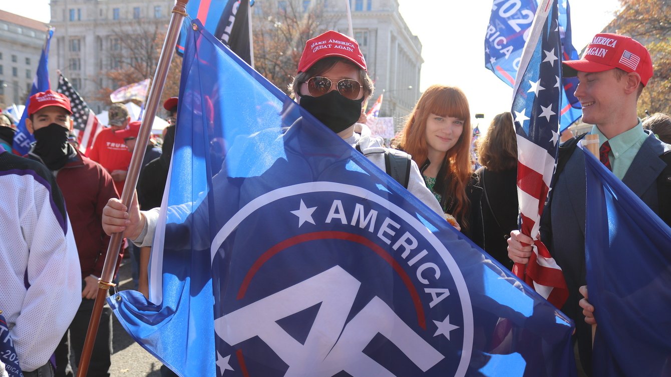 A man holding an "America First" flag, wearing a red MAGA hat, and a black face mask in a crowd of other right-wingers.
