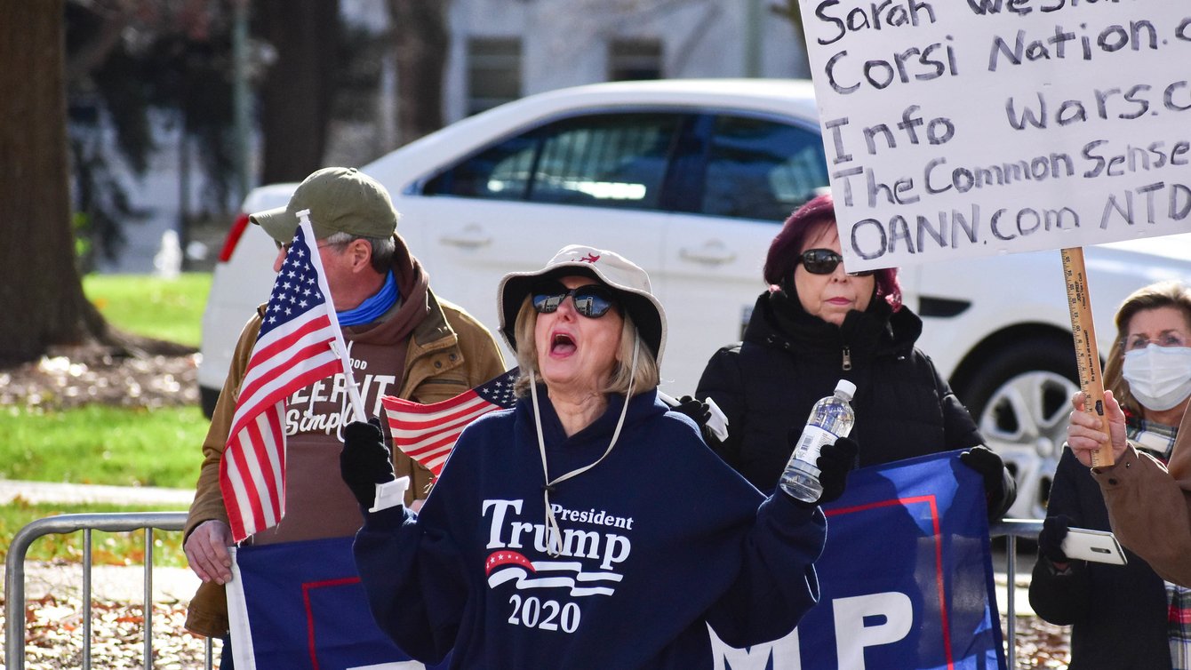 A White woman with blond hair, wearing a hat. She has a Trump 2020 sweatshirt on, and is holding an Amerian Flag and a water bottle. Her mouth is open like she's shouting something.