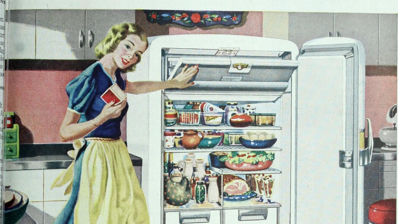 A white woman in a blue dress and a yellow apron, standing in front of an open fridge full of food.