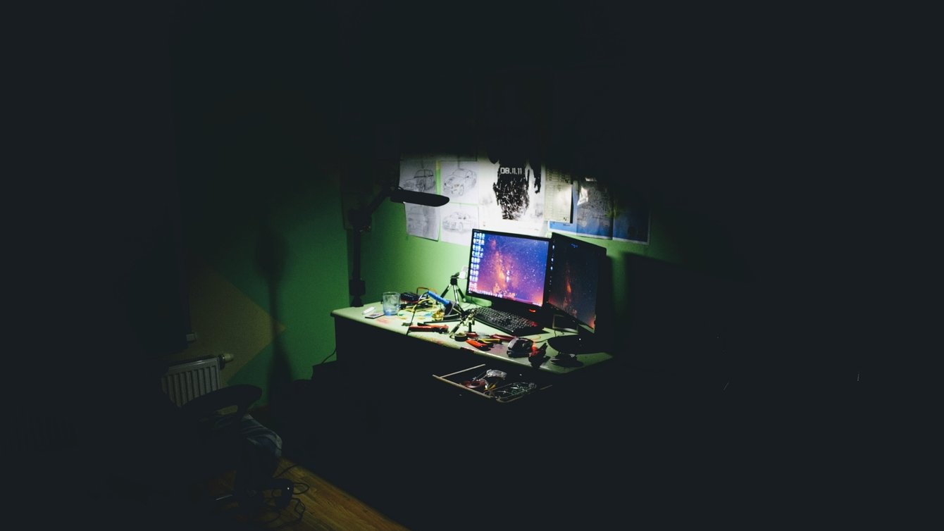 Two computer screens on a desk in a dark room. The light is focused on the computers