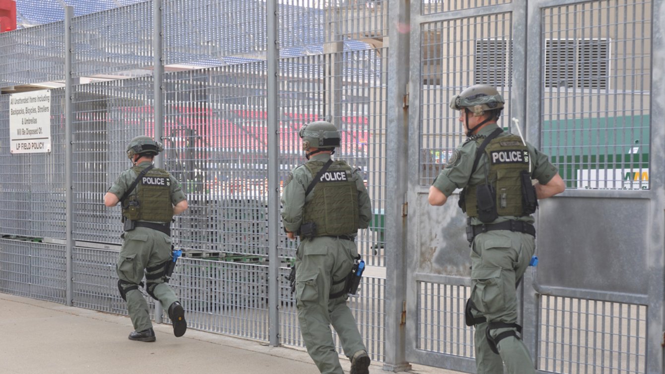 Three men in army training suits running along a large metal fence