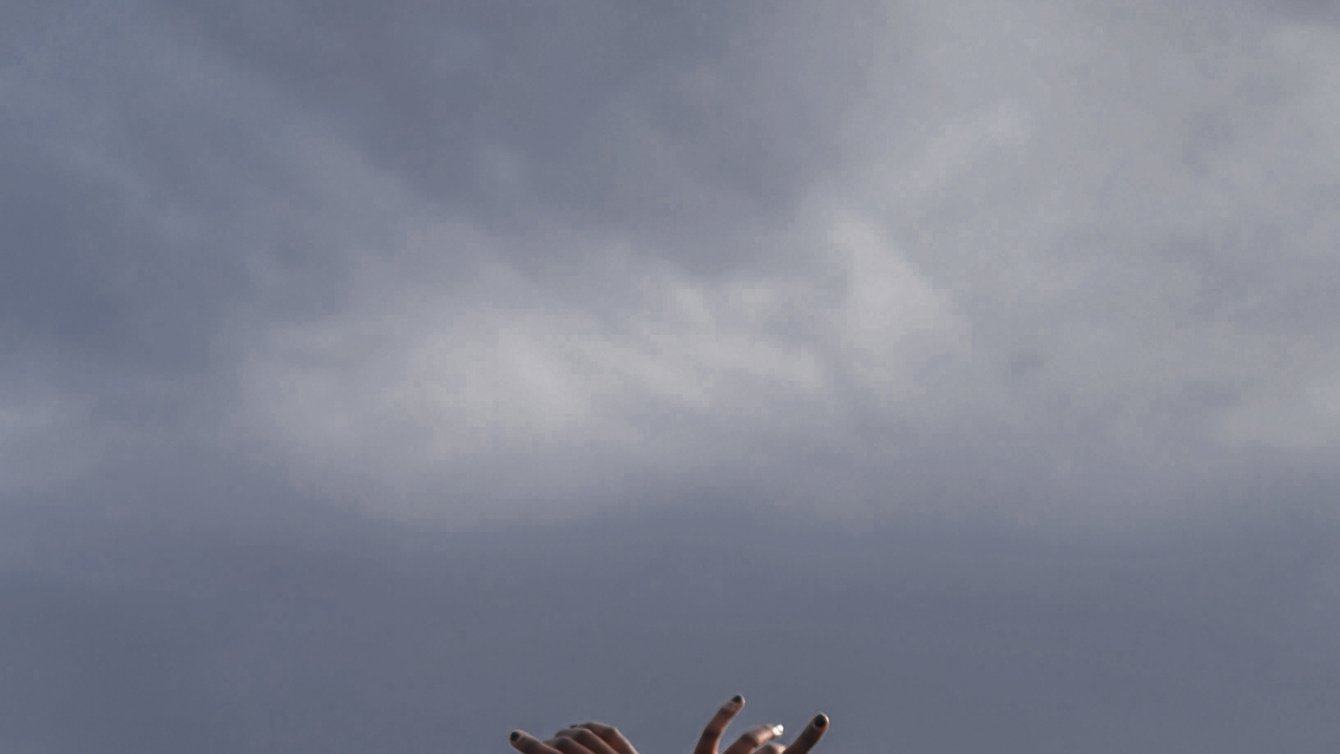 Black woman in field facing away from camera with arms in the air crossed at the wrist. 