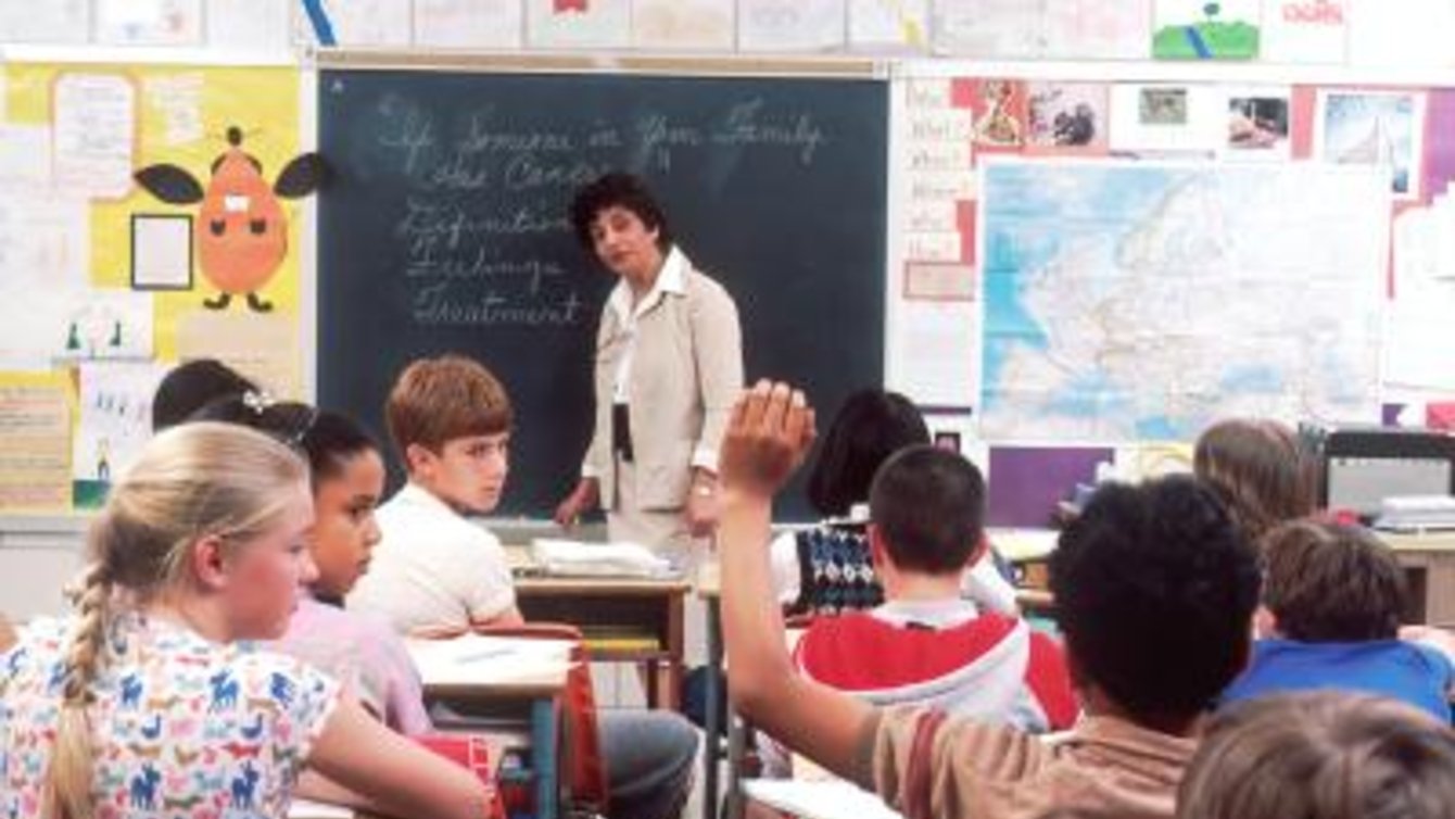 A teacher looking at a classroom of students, with one student's hand in the air