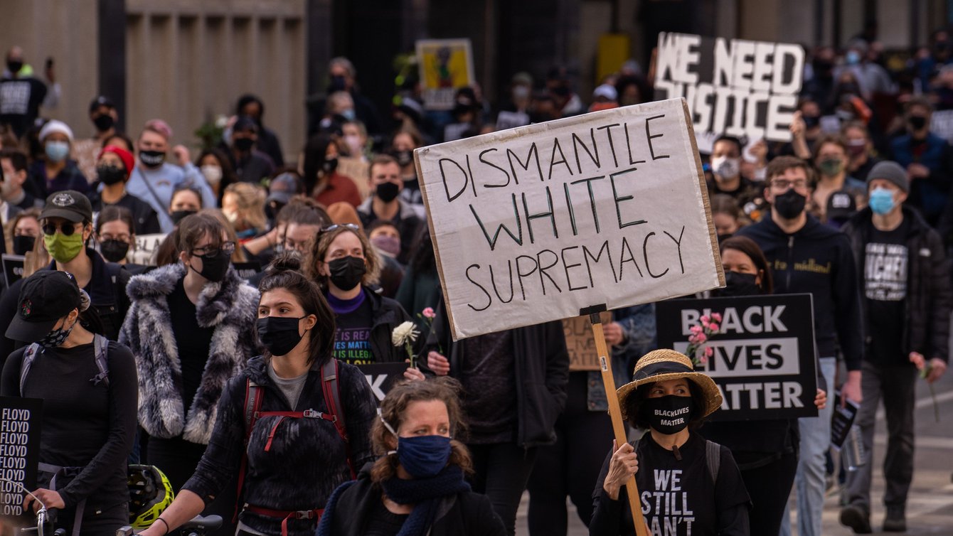 A person wearing a mask at a protest holding a sign that reads "Dismantle White Supremacy"