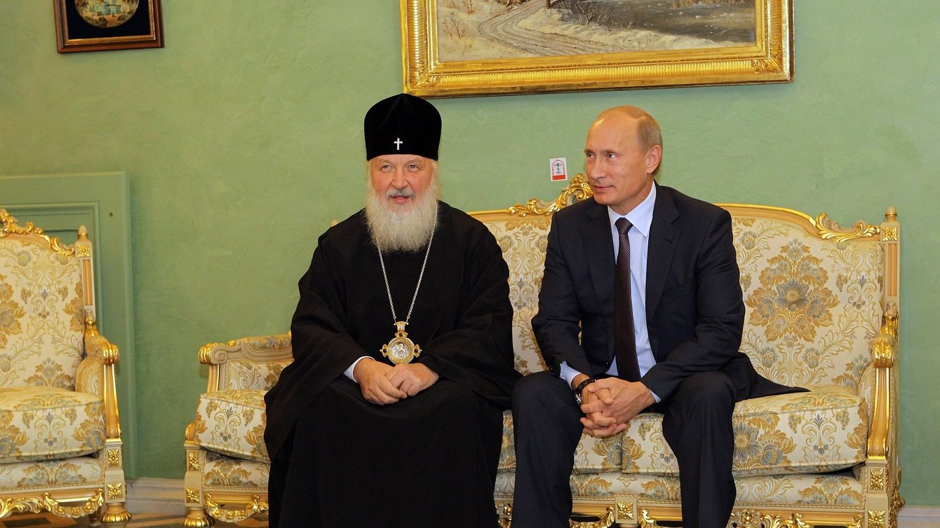 A man in a black robe and a black hate with a cross on it sitting next to Vladimir Putin on a golden couch.