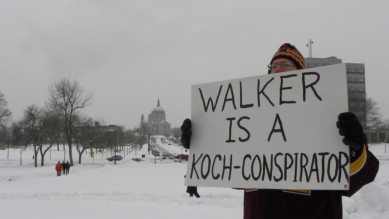 A man in winter clothes holding a board that says "walker is a koch-conspirator"
