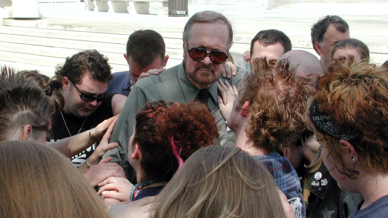 A white man in the center of a circle, with other people bowing their heads and placing a hand on his shoulder and arms.
