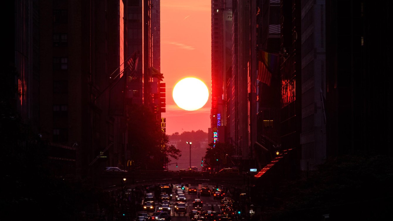 A sunset between buildings and a road full of vehicles.