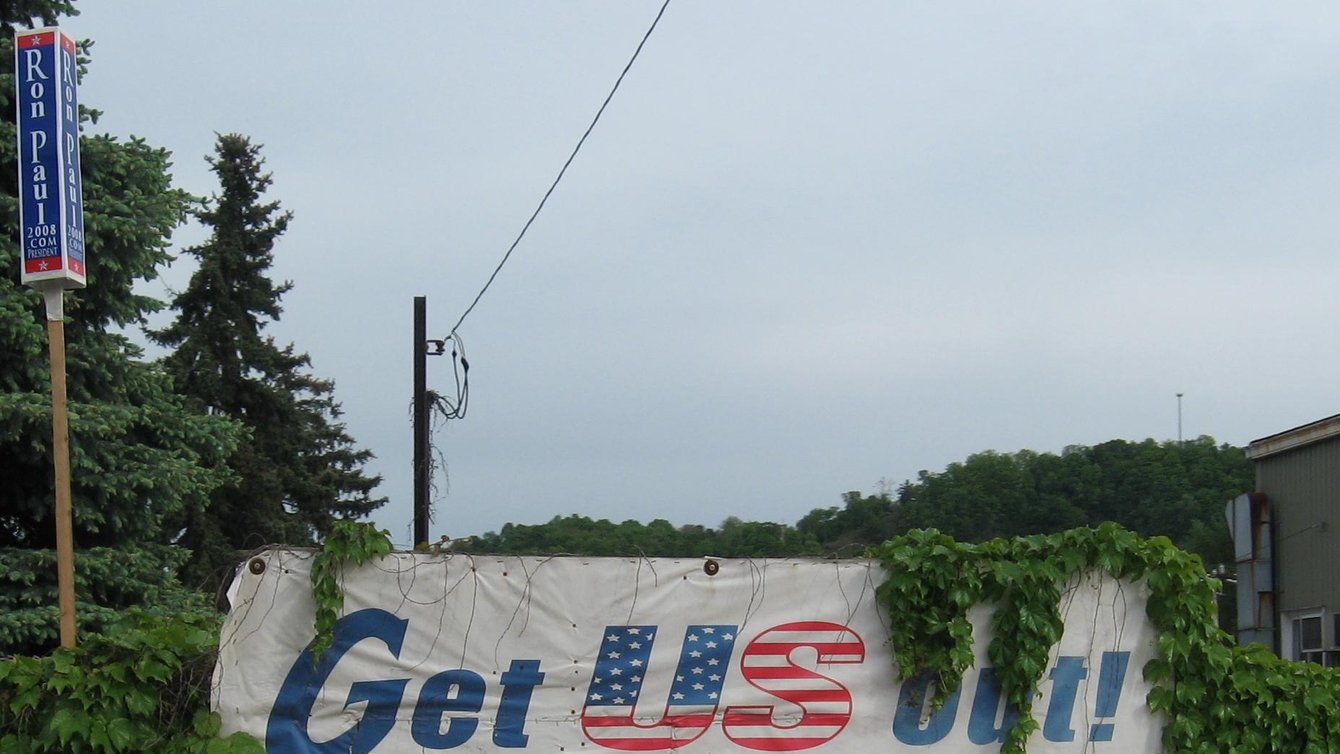 A sign covered by plants. The sign reads "Get US out! of the United Nations (888) JBS USA1"