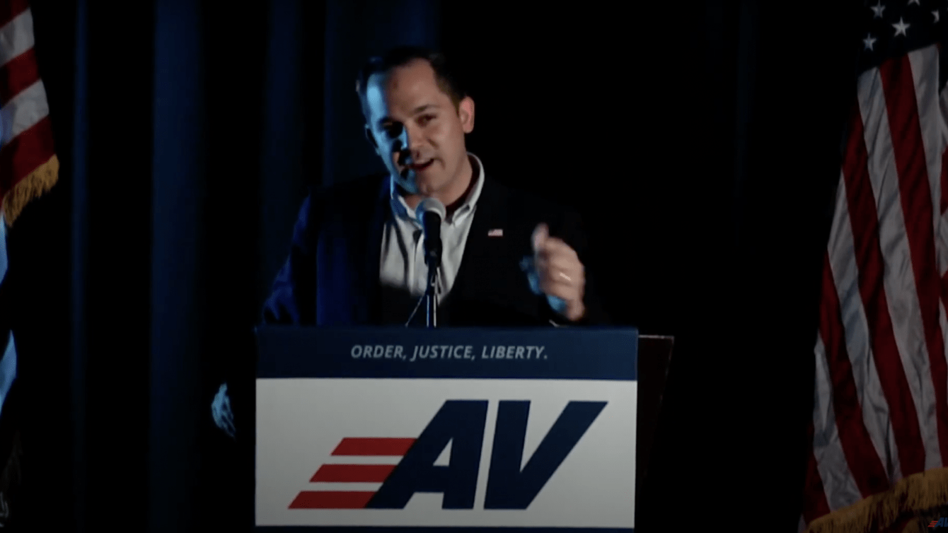 A man in blue suit standing at a podium with the letters AV on it speaking into a mic with two American flags behind him.