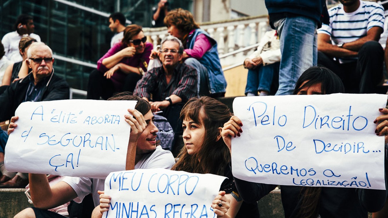 People sitting on stairs. Three people hold signs in Portugese, written with blue ink on white paper.