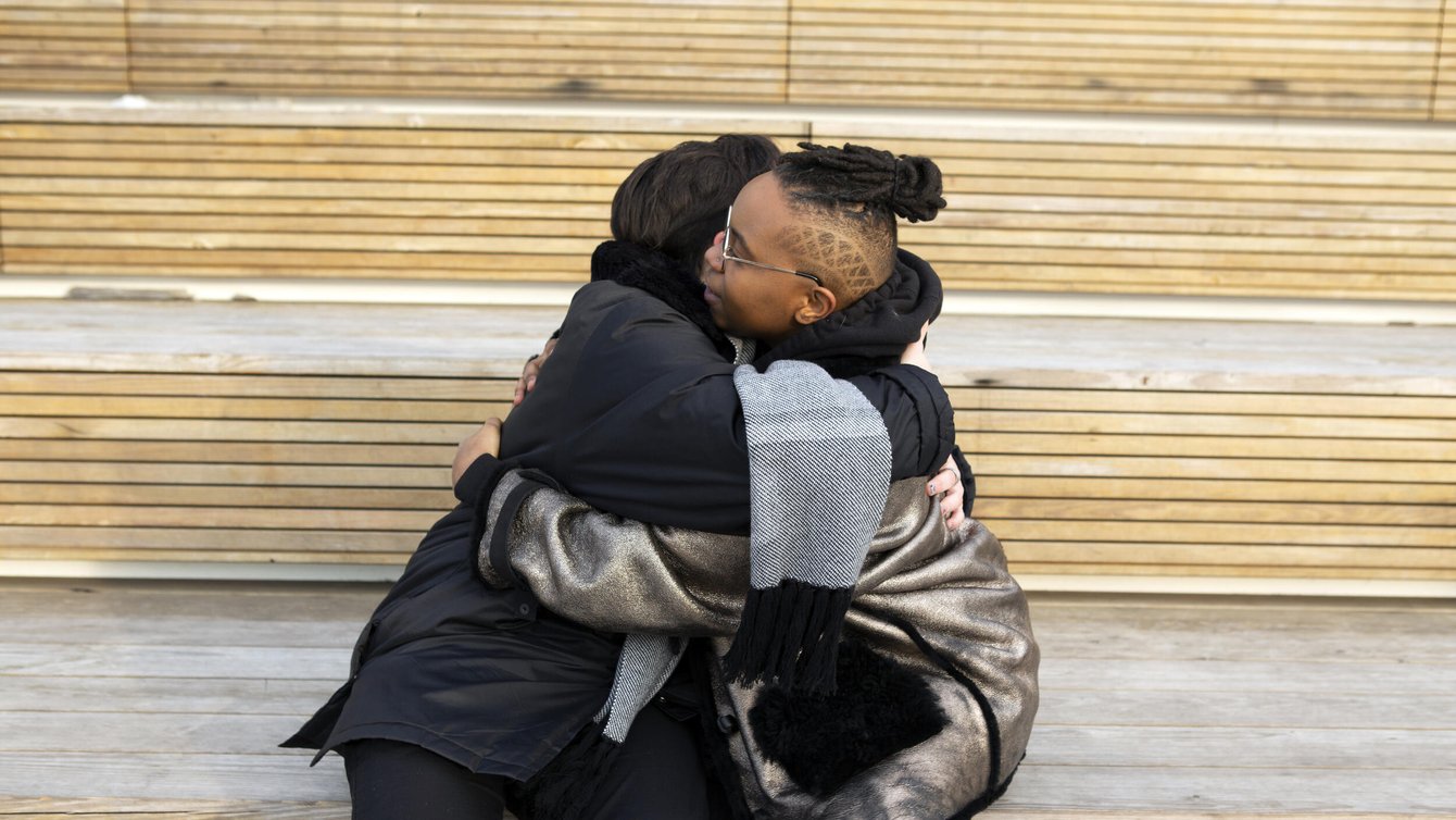 Two transmasculine people embracing while sitting on stairs.