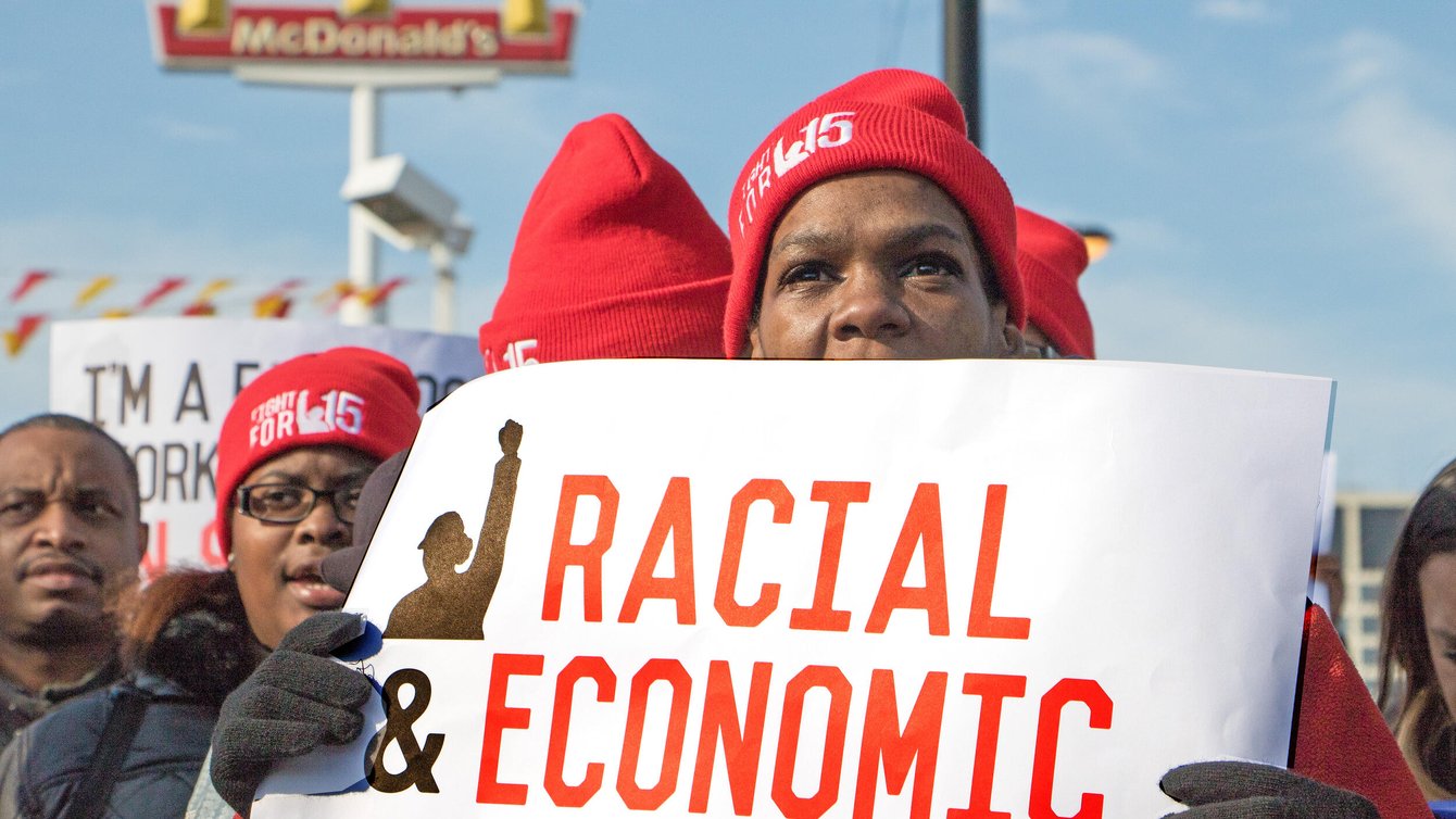 Woman in hat and gloves holds sign reading "Racial Economic Justice Now."