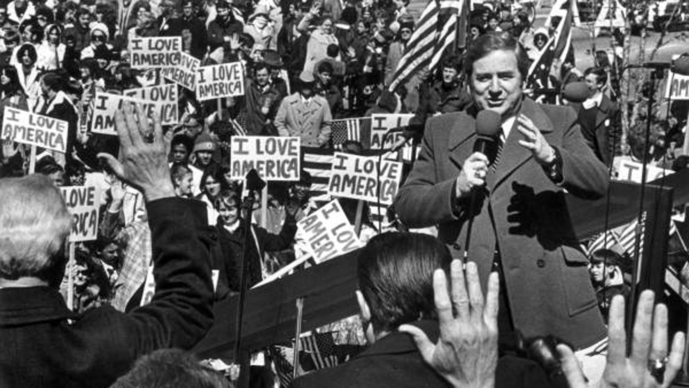 Jerry Falwell, a white man wearing a suit coat, speaks in front of a crowd holding signs reading "I love America."