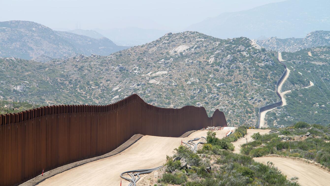 a brown fence following around a dirt road that fades into green mountains.