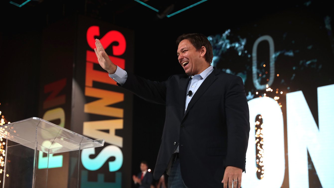 White man on stage with hand raised smiling, you can see the letters "ESANTIS" projected behind him.