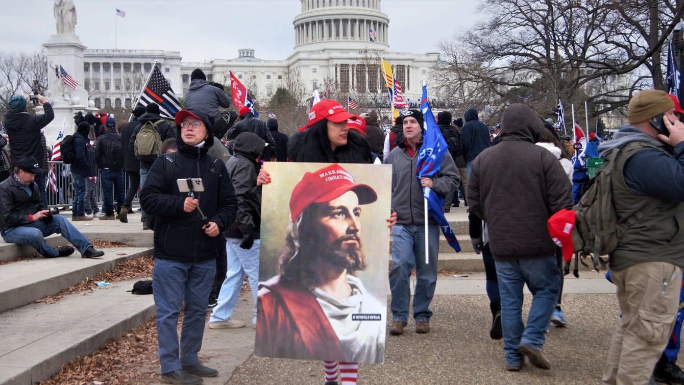 A woman wearing a Make America Great Again cap carrying an image of Jesus wearing a MAGA hat with "WWG1WGA" on his lapel