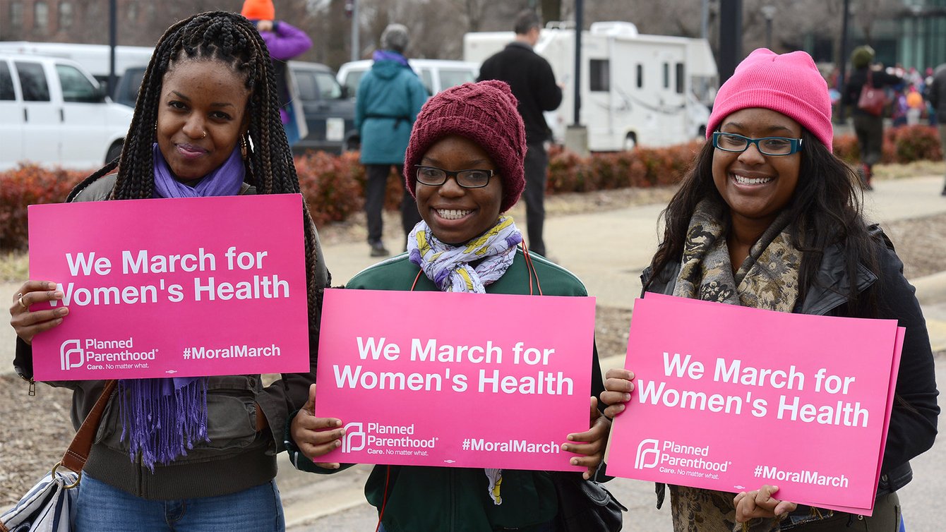 Three Black women wearing winter clothes holding signs that say "we march for Women's Health"