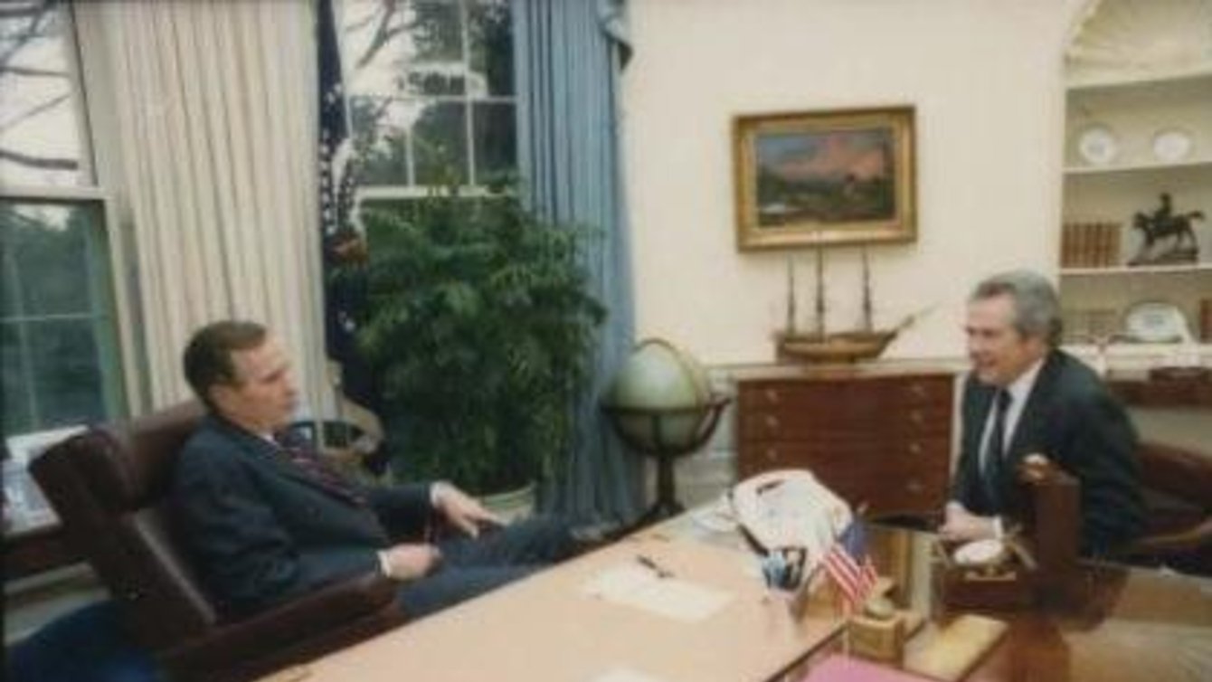 A man in a brown chair leaning back, and a man in leaning forward sitting opposite him.