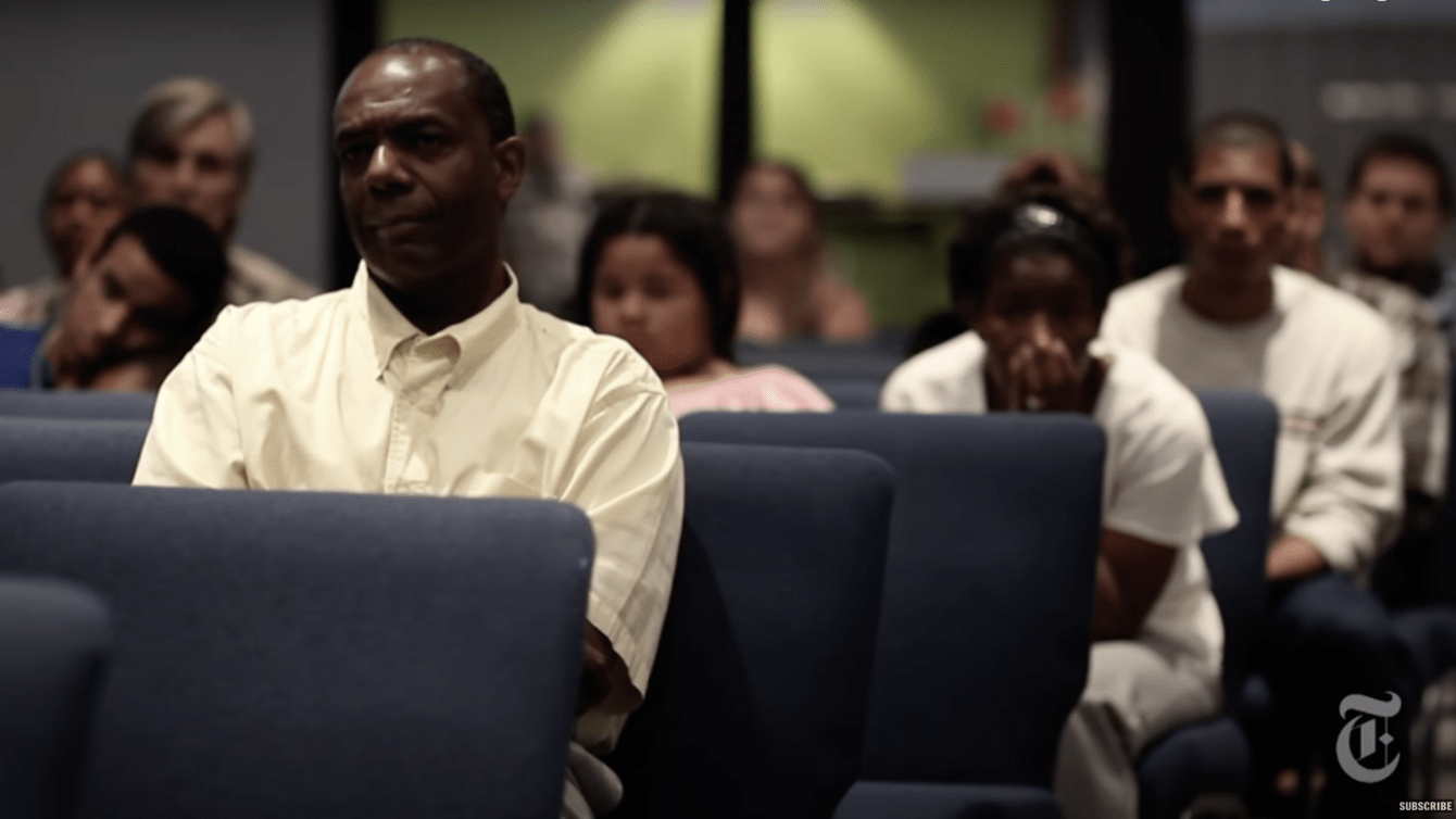 A black man in the foreground sitting in a grey chair, people behind him.