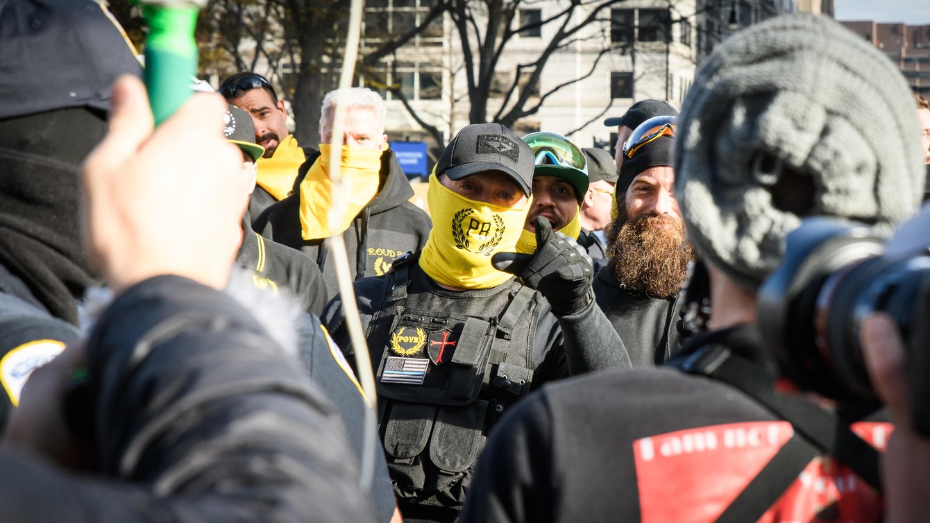 A Proud Boy with a neck warmer covering his mouth points mid-speech at the camera