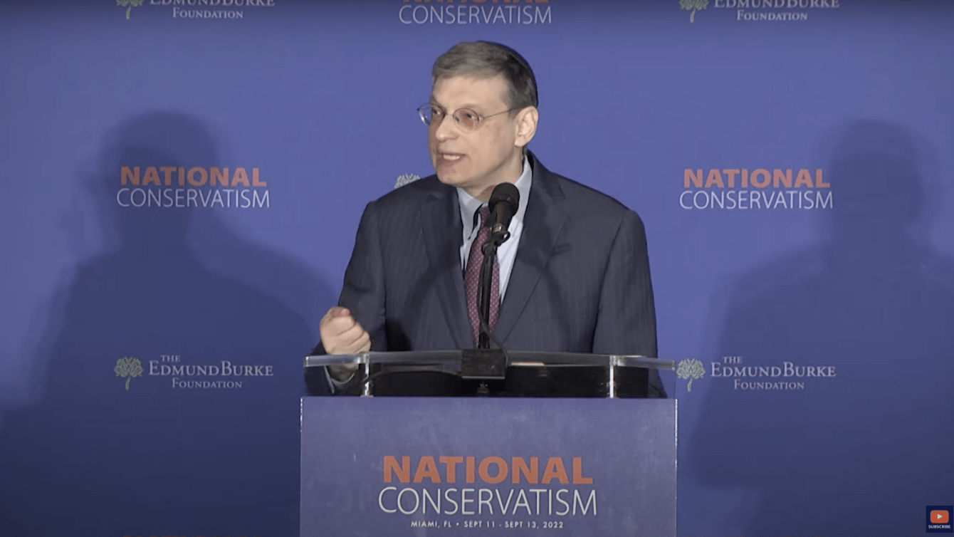 A Man in spectacles with greying hair talking at a podium that says "National Conservatism"