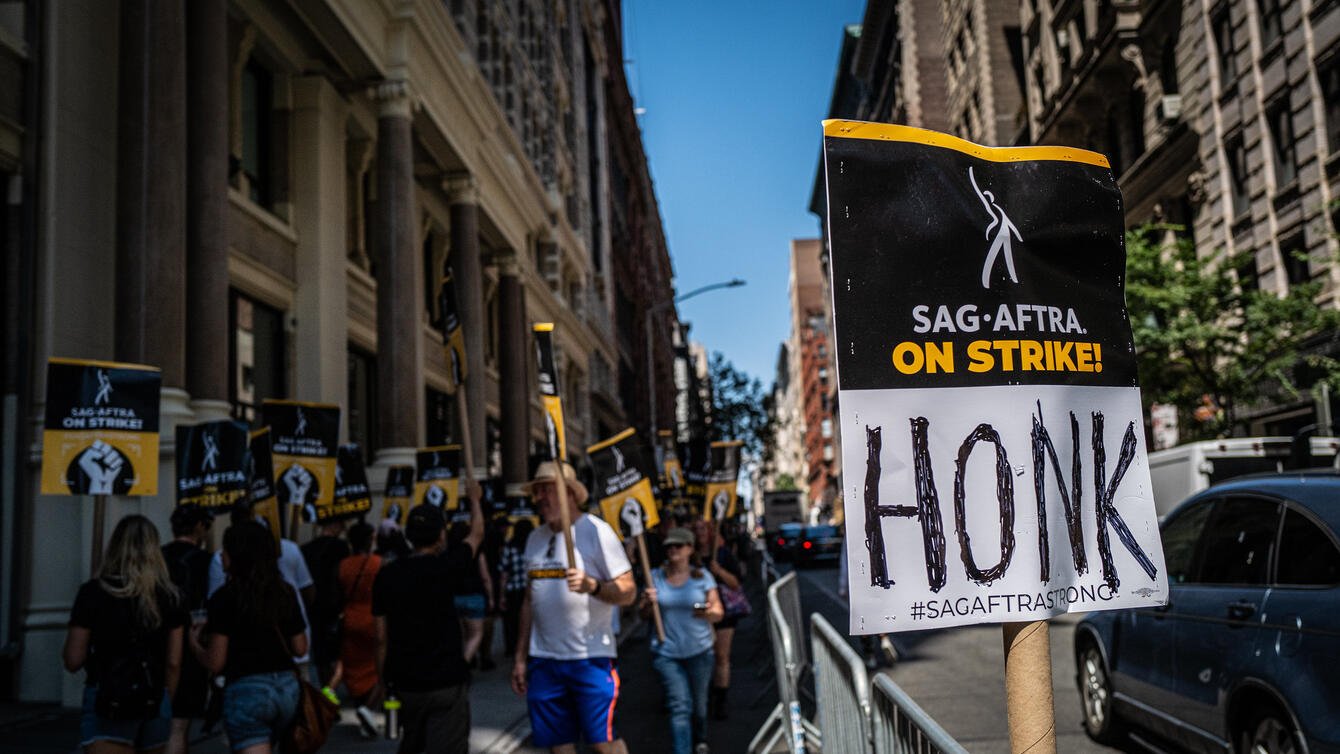 Protesters in the background. The Foreground has a sign that says "SAF-Aftra On Strike! Honk"