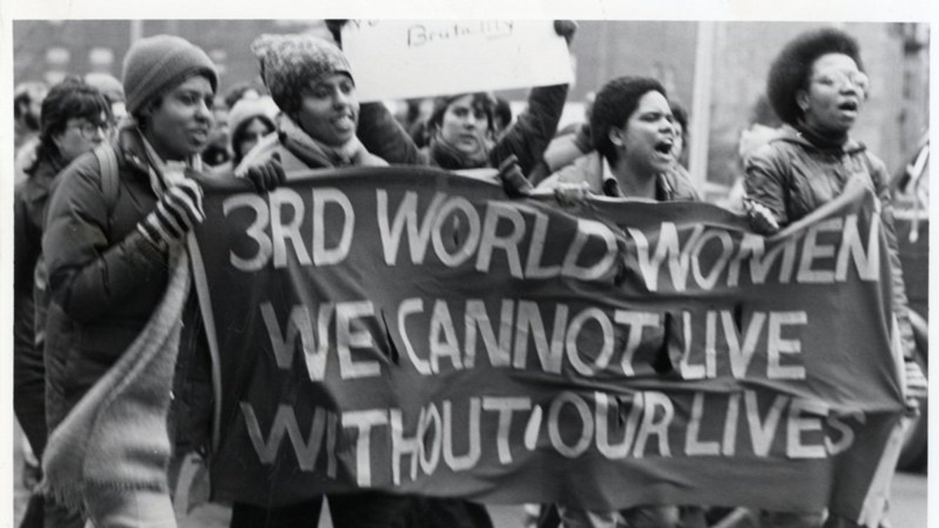 Four people walking holding a banner that says "3rd world women we cannot live without our lives"