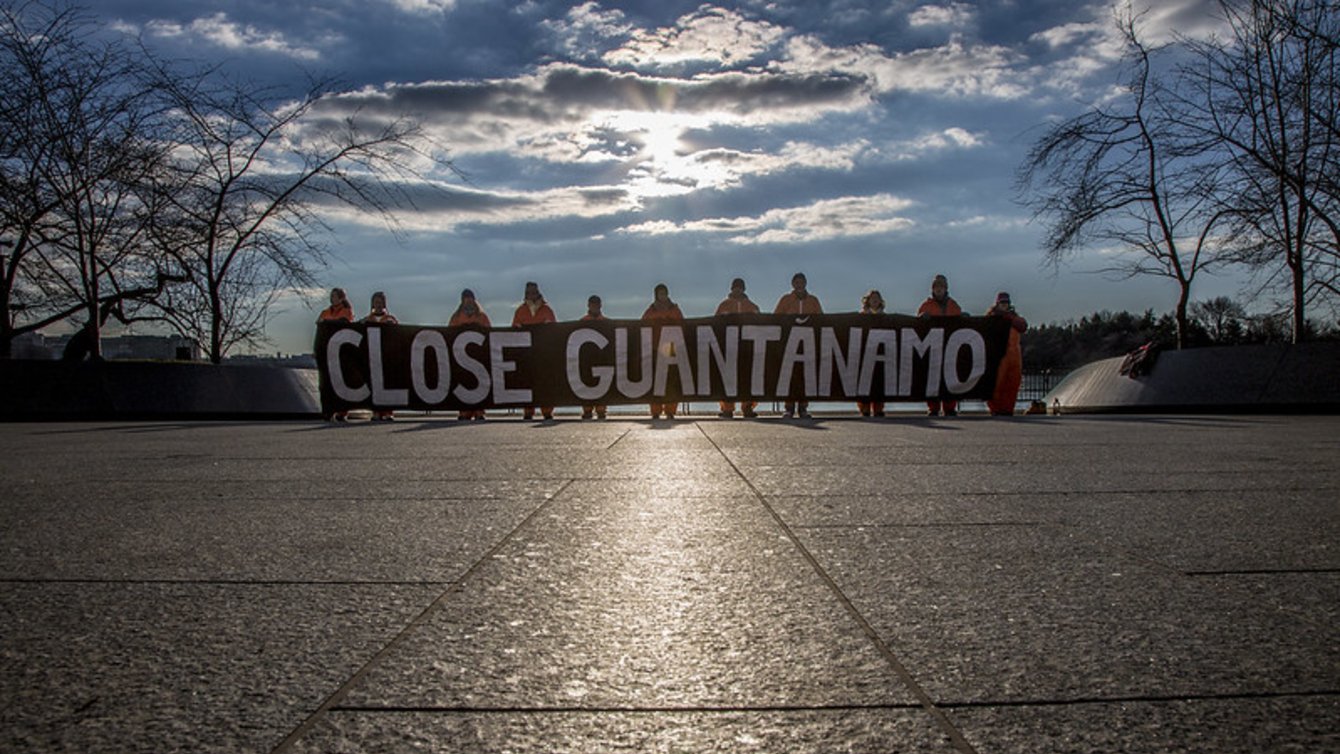 A row of people in orange jumpsuits holding a sign that says "close Guantanamo"