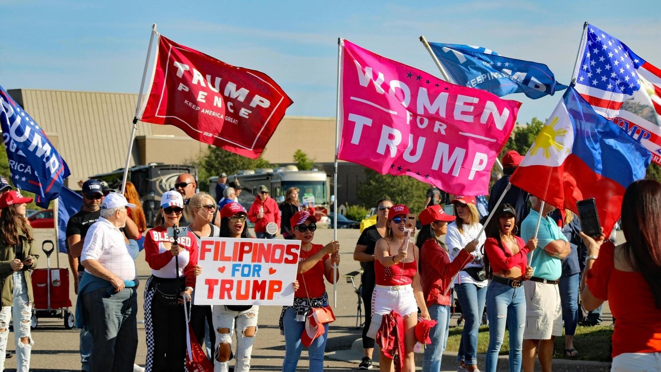 A group of women in red hats hold flags reading "Women for Trump," "Filipinos for Trump," and "Trump pence"