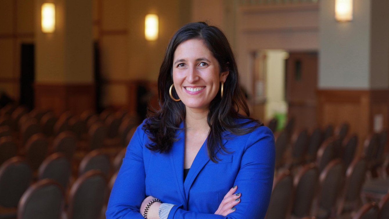 Smiling woman with long brown hair, gold hoop earrings, and a blue blazer, arms crossed