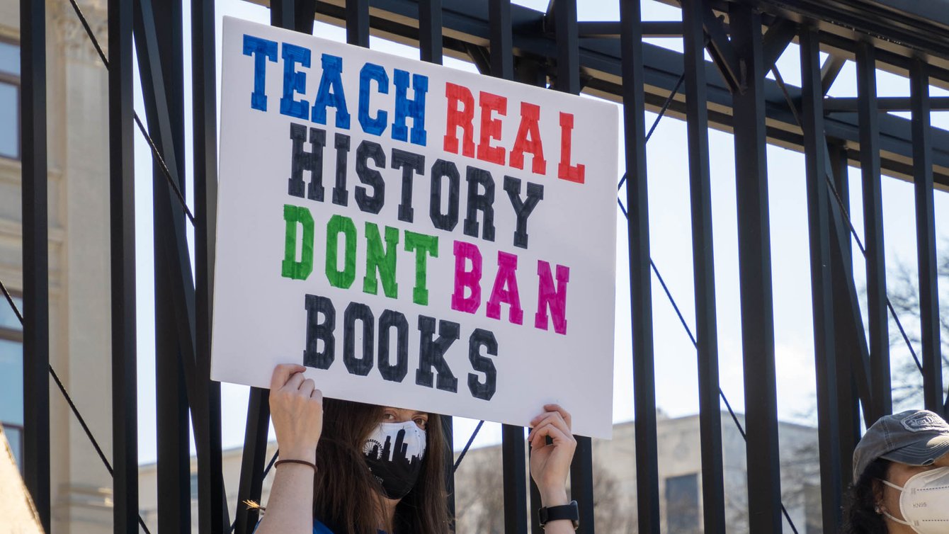 Woman in blue shirt holding a sign that reads "Teach real history don't ban books"