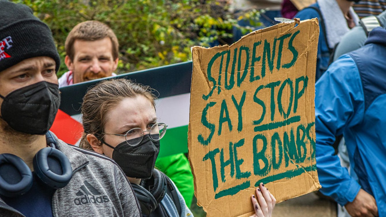 A person wearing a mask and glasses holds a sign reading "Students say stop the bombs"