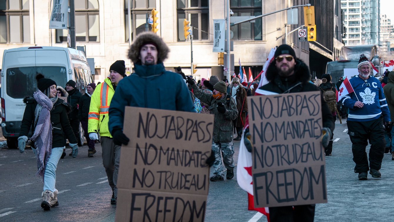 Two people in winter clothes holding signs that say "No jab pass, no mandates, no Trudeau, Freedom"