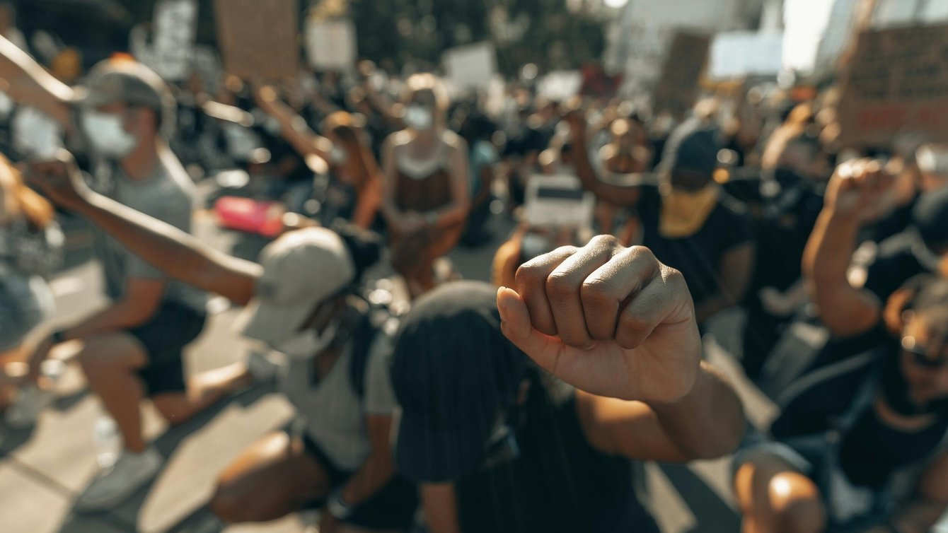 Focus on a raised fist, background of people kneeling in protest.