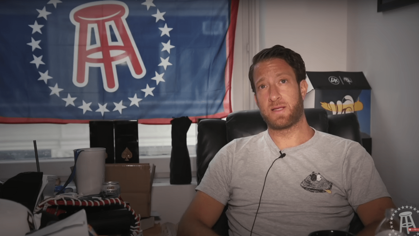A man in a grey T-shirt sitting in a chair with a flag behind him with the Barstool logo