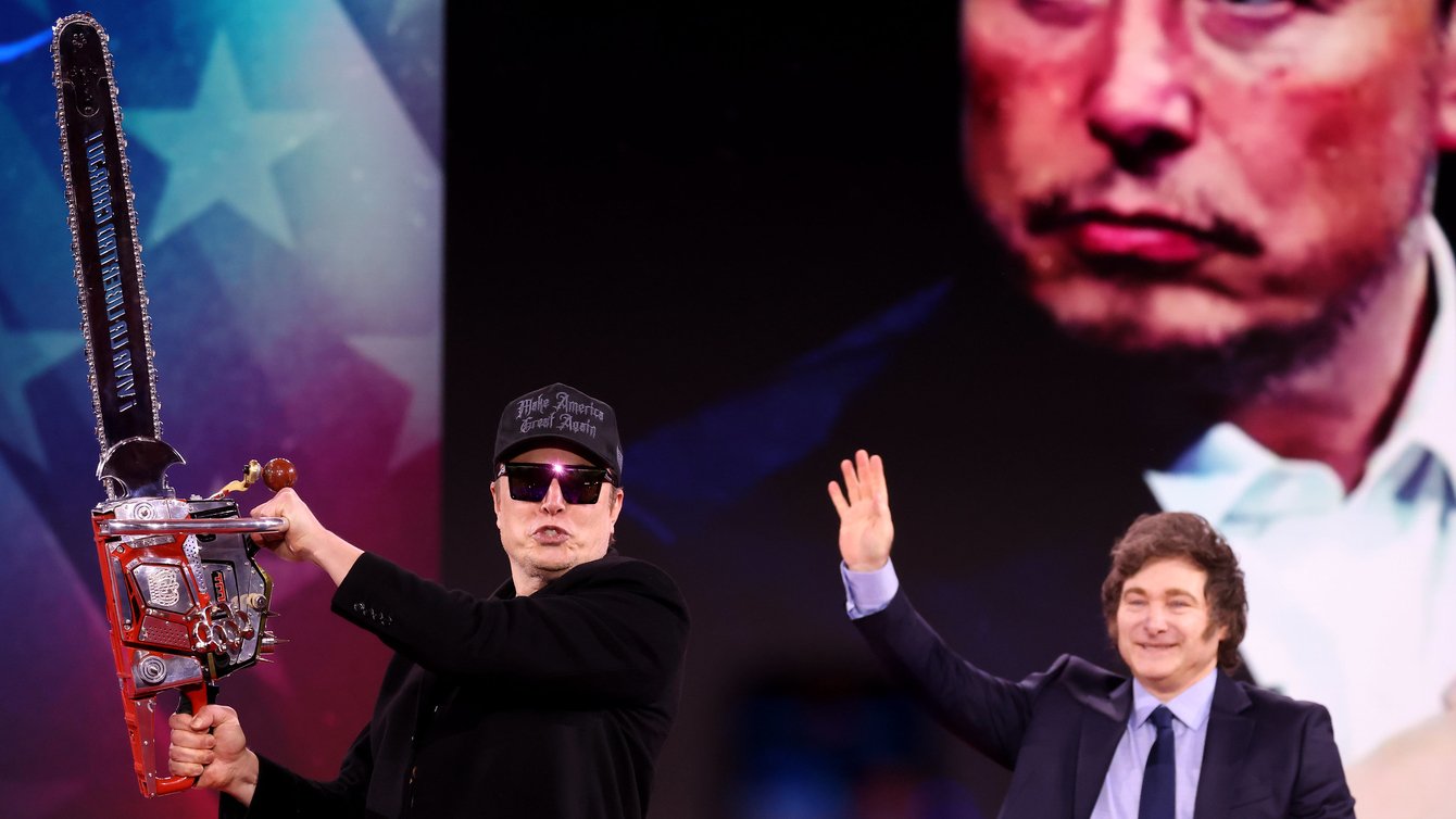 Two men standing in front of projector image. One man is waving at the crowd, the other man is holding a chainsaw