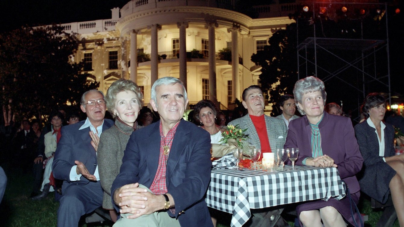 Six people sitting around a gingham table cloth on a picnic table with the white house in the background