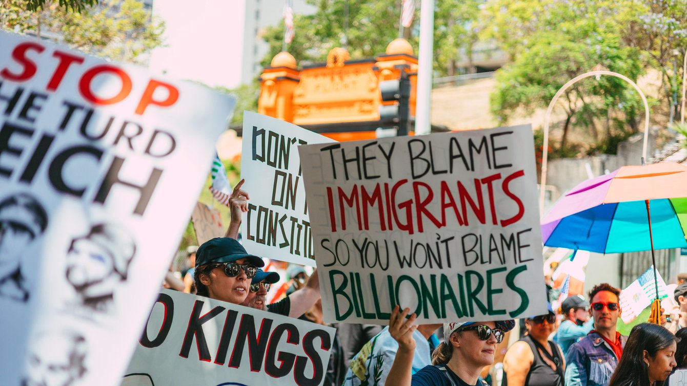 a person holding a sign that says "they blame the immigrants so you won't blame billionaires"