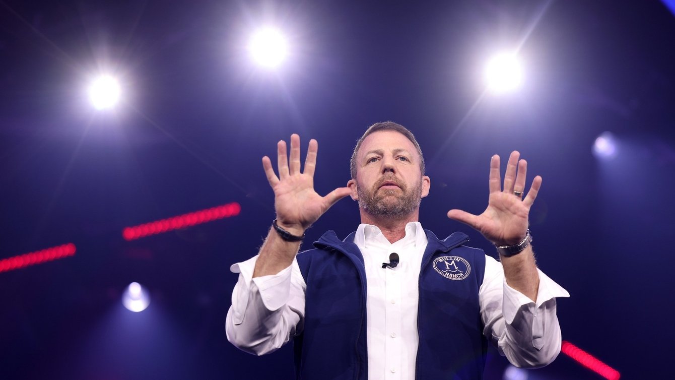 man in a white shirt and blue vest stands with hands raised with spotlights in the background
